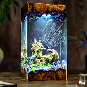 Puede incluir: Una pieza de arte de resina rectangular con una escena de dinosaurios en miniatura. La pieza tiene un marco de madera y un centro de resina transparente con un castillo, dos dinosaurios y patrones blancos arremolinados. El borde superior tiene un brillo azul y morado.