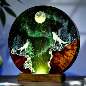 May include: A round, illuminated resin art piece featuring a nighttime forest scene with two white wolves howling at the moon. The artwork includes a green aurora borealis effect, trees, and a wooden base. The piece is a unique decorative item.