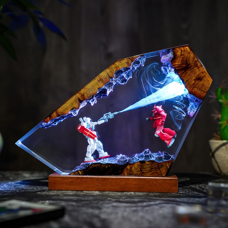 May include: A unique resin art piece featuring two robot figures in a battle scene. The artwork is encased in a clear resin with a wooden frame and base. The robots are white and red, with a blue energy beam. The piece is illuminated with blue light.