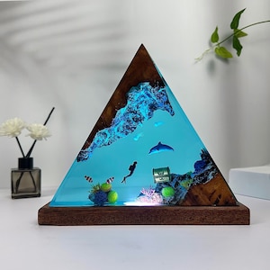 May include: A triangular resin sculpture depicting a blue and green ocean scene. The sculpture features a dolphin, a diver, and coral. The base is made of dark wood.