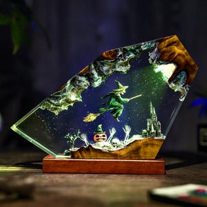May include: A Halloween-themed resin art piece featuring a green witch flying on a broomstick over a spooky landscape with a pumpkin, skeletons, and a castle. The artwork is set on a wooden base and has a light shining through it.