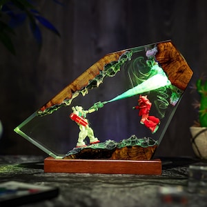 May include: A unique resin art piece featuring two robot figures engaged in battle. The figures are set within a clear resin block with a wooden border. The scene is illuminated with green light, creating a dynamic and artistic display. The base is made of wood.