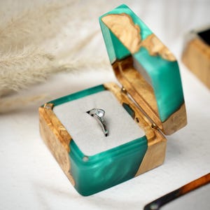 May include: A silver ring displayed in an open wooden ring box. The box features a teal resin inlay and light-coloured wood. The ring has a clear gemstone set on a silver band. The box is lined with grey fabric.