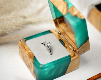 May include: A silver ring displayed in an open wooden ring box. The box features a teal resin inlay and light-coloured wood. The ring has a clear gemstone set on a silver band. The box is lined with grey fabric.