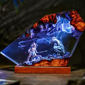 Può includere: Una scultura in resina raffigurante due figure di Predator in una scena di combattimento, racchiuse in un blocco trasparente e angolato. La scultura poggia su una base di legno ed è illuminata con tonalità blu e arancioni, creando un effetto drammatico. Le figure sono dettagliate e la resina ha un aspetto fumoso.