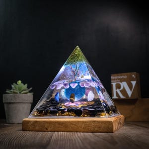 Pode incluir: Lâmpada de resina em forma de pirâmide com base de madeira. A lâmpada apresenta um interior azul e roxo com um design de árvore, rodeado por pedras pretas e um topo verde. A lâmpada tem cerca de 15 cm de altura.