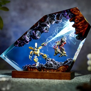 May include: A unique resin art piece featuring two robot figures in a dynamic battle scene. The artwork is encased in a clear, angled resin block with a blue background, and a wooden base. The robots are yellow and red, with a beam of light.