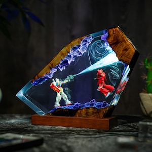 May include: A unique resin art piece featuring two robot figures in a battle scene. The artwork is encased in a clear resin block with a wooden frame and base. The figures are white, red, and blue, with a blue energy beam.