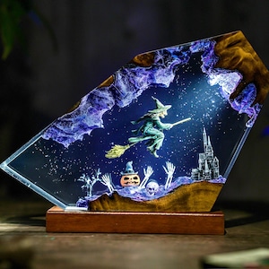 May include: A Halloween-themed resin art piece featuring a witch on a broomstick flying across a starry night sky. The artwork includes a castle, a pumpkin, and skeletal figures. The piece is set on a wooden base.