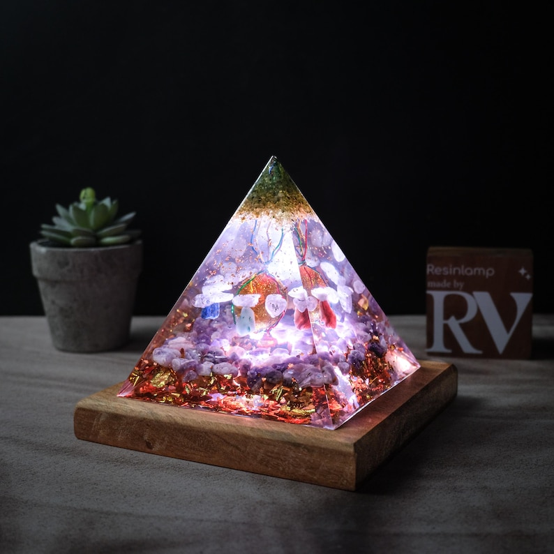 May include: A pyramid-shaped resin lamp with a wooden base. The lamp is filled with crystals and gold flakes, and it glows with a soft, white light. The text "Resinlamp made by RV" is visible on a small card in the background.