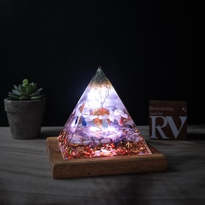 May include: A clear pyramid-shaped resin lamp with embedded crystals and gold flakes, lit from within, on a wooden base. The lamp is on a dark surface with a small potted plant in the background. The text "Resinlamp made by RV" is visible on a card in the background.