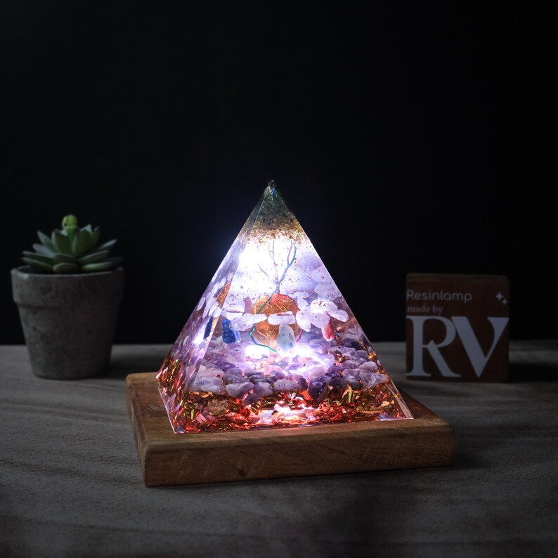 May include: A clear pyramid-shaped resin lamp with embedded crystals and gold flakes, lit from within, on a wooden base. The lamp is on a dark surface with a small potted plant in the background. The text "Resinlamp made by RV" is visible on a card in the background.
