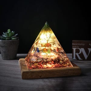 May include: A clear glass pyramid filled with colorful crystals and stones, lit from within, sits on a wooden base. The pyramid is a resin lamp, made by RV.