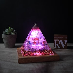 May include: A pink and purple resin pyramid with crystals and gold glitter inside, lit from within, on a wooden base. The pyramid is on a wooden table with a small succulent plant in a pot to the left. A small sign with the text "Resinlamp made by RV" is to the right.