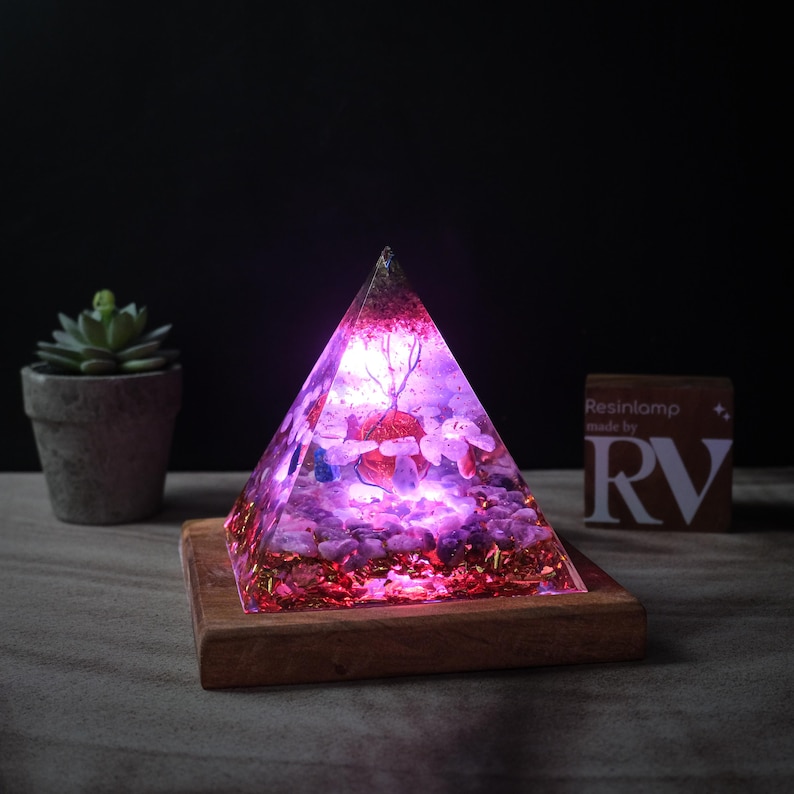 May include: A pink and purple resin pyramid with crystals and gold glitter inside, lit from within, on a wooden base. The pyramid is on a wooden table with a small succulent plant in a pot to the left. A small sign with the text "Resinlamp made by RV" is to the right.