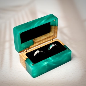 May include: Open teal and wood ring box with two rings inside. The box is rectangular with a hinged lid and a black velvet interior. The rings are silver with clear stones. The box is on a white surface.