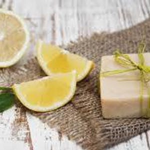 Può includere: Natura morta con mezzo limone, due spicchi di limone, una foglia verde e una saponetta avvolta con un nastro verde. Gli elementi sono disposti su un tessuto di juta e una superficie di legno verniciata di bianco.