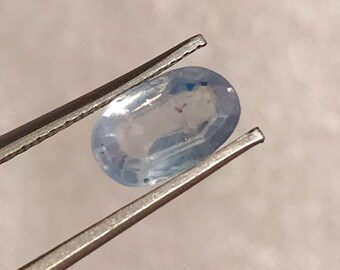 Könnte beinhalten: Ein hellblauer, oval geschliffener Edelstein, der von einer Metallpinzette gehalten wird. Der Stein ist transparent mit feinen Einschlüssen. Die Pinzette ist abgewinkelt, um die Form und Facetten des Steins hervorzuheben. Der Hintergrund ist neutral.