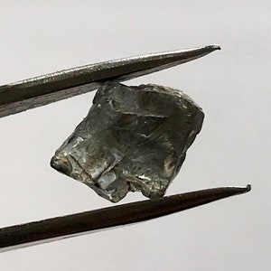 May include: A grey, faceted gemstone held by tweezers. The gemstone has a rough, natural appearance.