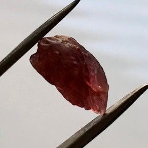 May include: A raw, pink, rough-cut gemstone held by tweezers. The gemstone is translucent and has a slightly irregular shape.