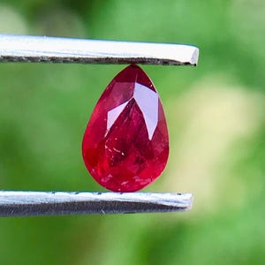 Op de afbeelding: Een gefacetteerde, peervormige robijn, vastgehouden door een pincet. De robijn heeft een diepe, levendige rode kleur, met meerdere facetten die het licht vangen. De achtergrond is een zacht, wazig groen, wat een contrast vormt met de edelsteen.