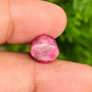 Puede incluir: Una pequeña gema rosa pulida con forma redondeada. La piedra tiene una calidad translúcida con sutiles patrones internos y variaciones de color. La gema se sostiene entre dos dedos sobre un fondo verde borroso.