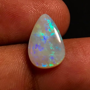 May include: A pear-shaped opal gemstone with a white base color and iridescent flashes of blue, green, and yellow. The polished stone is held against a dark background, showcasing its vibrant play-of-color.