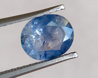 Könnte beinhalten: Ein facettierter, ovalförmiger blauer Edelstein wird von einer silbernen Pinzette vor einem weißen Hintergrund gehalten. Der Stein weist verschiedene Blautöne und interne Einschlüsse auf, die durch die Facetten sichtbar werden.