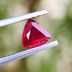 Op de afbeelding: Een driehoekige, gefacetteerde, dieprode edelsteen, vastgehouden door een zilveren pincet. De edelsteen heeft een gepolijst oppervlak dat licht reflecteert en zijn levendige kleur laat zien. De achtergrond is een zacht, wazig groen.