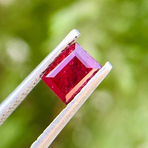 Può includere: Una pietra preziosa di rubino rosso intenso con taglio rettangolare, tenuta in una pinzetta su uno sfondo verde sfocato.
