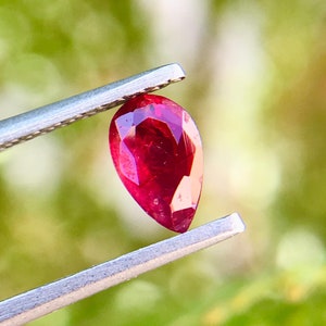 Op de afbeelding: Een peervormige robijn edelsteen vastgehouden door een pincet. De robijn is diep rood van kleur en heeft een briljante glans.