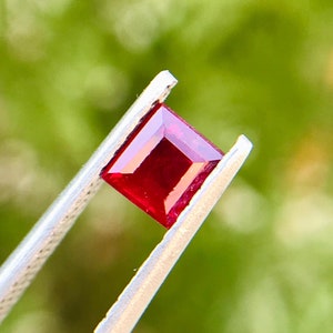 Peut inclure: Une pierre précieuse carrée rouge foncé tenue dans des pinces sur un fond vert.