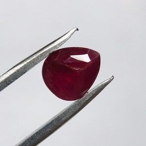 Puede incluir: Una piedra preciosa de color rojo intenso en forma de pera sostenida por unas pinzas.