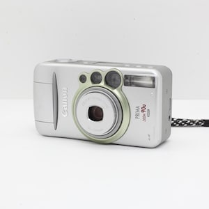 Könnte beinhalten: Silberne Canon Prima Zoom 90u Kamera mit einer schwarz-weißen Handschlaufe. Die Kamera hat ein Objektiv mit grünem Ring und das Canon-Logo auf der Vorderseite. Auf weißem Untergrund.