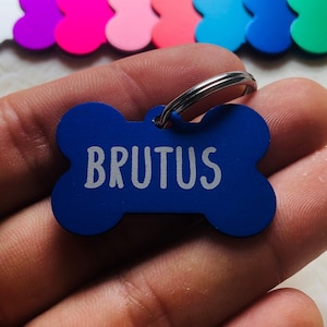 Può includere: Medaglietta per cani a forma di osso blu con un anello argentato e il nome "BRUTUS" inciso in lettere bianche.