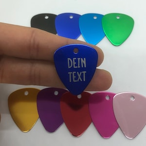 Puede incluir: Un conjunto de diez púas de guitarra en varios colores, incluyendo negro, azul, verde, dorado, morado, rojo, rosa y azul claro. Una púa es azul con el texto "DEIN TEXT" grabado en ella.