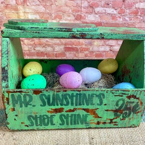 May include: A vintage green wooden shoe shine box with the words "Mr. Sunshine's Shoe Shine 25¢" painted on the front. The box is filled with colorful Easter eggs and straw.