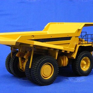 Pu&ograve; includere: Un camion ribaltabile giocattolo giallo con una griglia nera e una gabbia nera sul retro. Il camion ha grandi pneumatici neri ed &egrave; su uno sfondo blu.