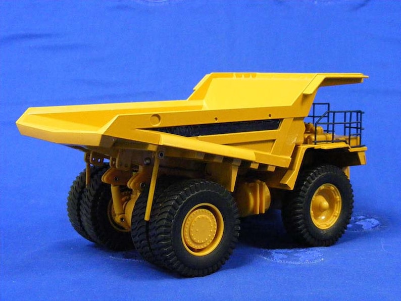 Pu&ograve; includere: Un camion ribaltabile giocattolo giallo con una griglia nera e una gabbia nera sul retro. Il camion ha grandi pneumatici neri ed &egrave; su uno sfondo blu.