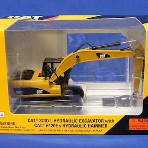 May include: A yellow and black 1:50 scale die-cast model of a CAT 323D L Hydraulic Excavator with a CAT H120E s Hydraulic Hammer. The model is in a clear plastic box with a yellow background. The box has a warning label that says "Choking Hazard - Small parts. Not for children under 3 years."