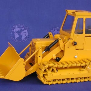 Op de afbeelding: Een gele Caterpillar 941 model bulldozer met een voorste bak en rupsbanden. Het model heeft een cabine met ramen en het Caterpillar-logo aan de zijkant. De bulldozer staat tegen een blauwe achtergrond.