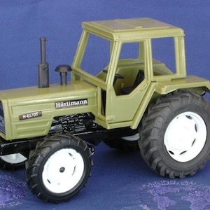 Pode incluir: Um trator de brinquedo verde com um tubo de escape preto e um volante preto. O trator tem um rótulo branco H-6170T na frente e um rótulo Hürlimann no lado.