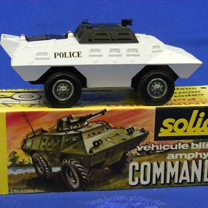 Puede incluir: Un vehículo de policía de juguete blanco con detalles negros y la palabra "POLICE" en el lateral. El vehículo está sentado en una caja amarilla con la palabra "solido" y el texto "véhicule blindé amphybie COMMANDO" en blanco y negro.