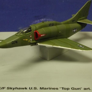 Peut inclure: Un modèle d'avion A4 E/F Skyhawk vert et blanc avec le texte "VF101" et "074" sur le côté. Le modèle est sur une base blanche et porte le texte "A4 E/F Skyhawk U.S. Marines "Top Gun" art. 5223" sur la base.