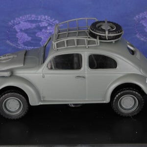 May include: A gray toy model of a Volkswagen Beetle with a roof rack and a spare tire mounted on the back. The car has a detailed interior and exterior, including a spare tire on the roof rack.