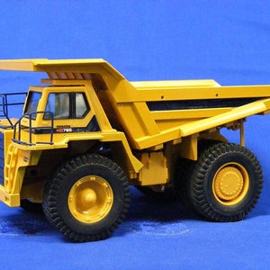 Pu&ograve; includere: Autocarro ribaltabile giocattolo giallo con dettagli neri e una gabbia di sicurezza nera. Il camion ha un cassone grande e quattro pneumatici grandi. Il camion ha il testo "KOMATSU HD785" sul lato.