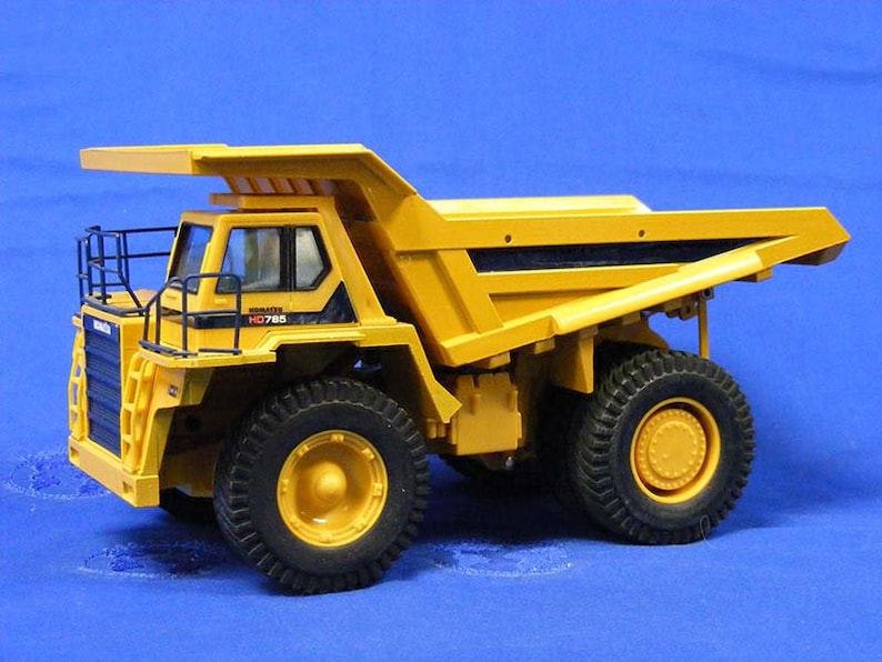 Pu&ograve; includere: Autocarro ribaltabile giocattolo giallo con dettagli neri e una gabbia di sicurezza nera. Il camion ha un cassone grande e quattro pneumatici grandi. Il camion ha il testo "KOMATSU HD785" sul lato.