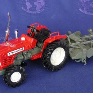 May include: A red toy tractor with a silver exhaust pipe and a green plow attachment. The tractor has the text "イセキ トラクター 509" printed on the side.