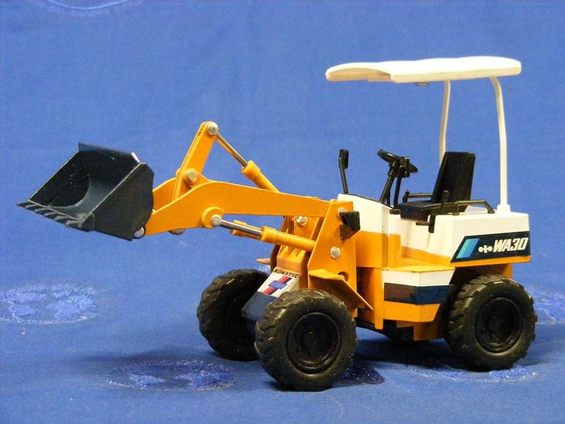 May include: A yellow and white toy backhoe with a black bucket. The backhoe has a white canopy and the text "WA30" on the side.