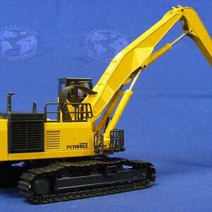 May include: Yellow and black toy model of a Komatsu PC1100LC excavator with a magnetic grapple. The excavator has a long yellow arm with a black chain and a round magnetic plate at the end.